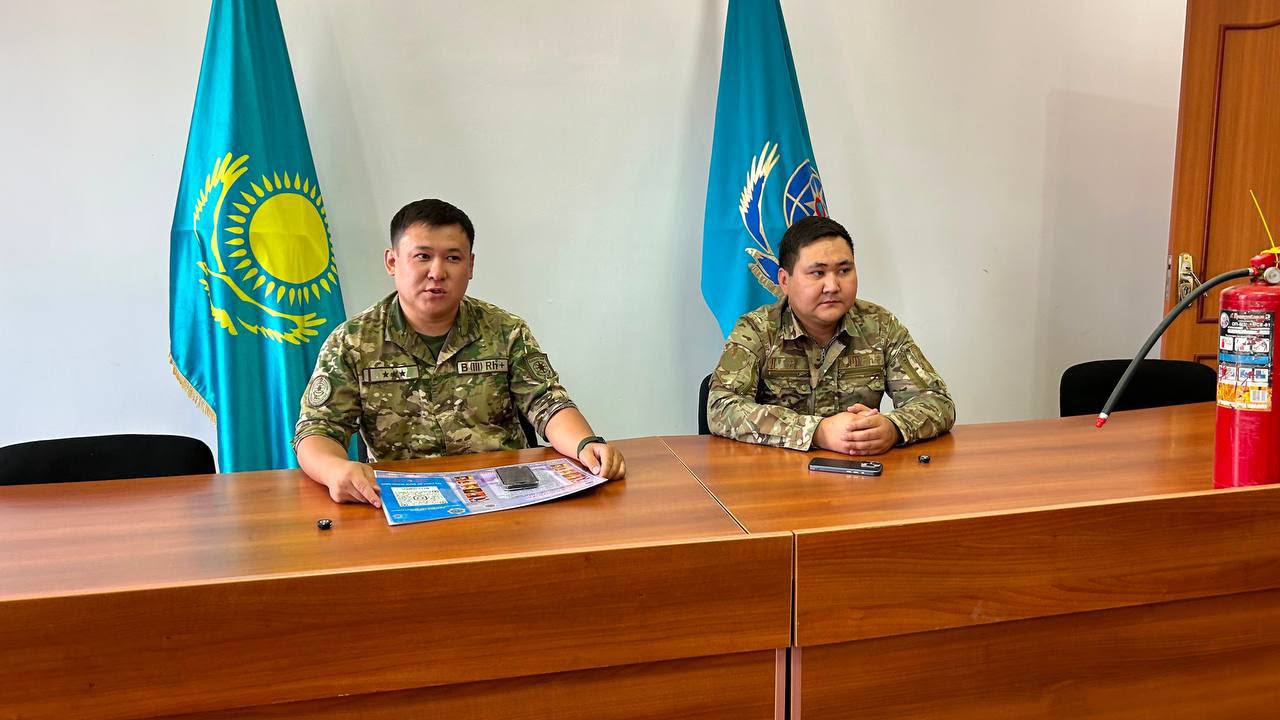 Қазақстан туларының алдында үстел басында отырған әскери формадағы екі ер адам.