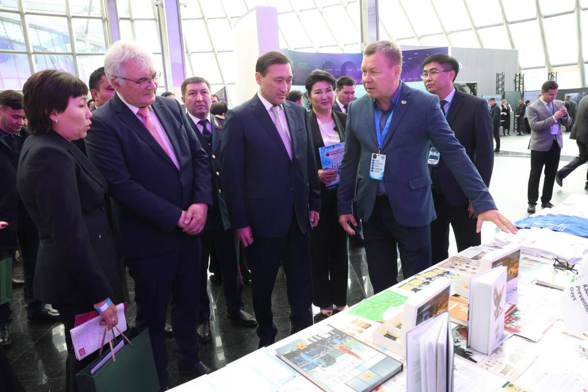 Ресми киінген адамдар тобы көрмедегі кітаптар мен жарнамалық материалдарды қарап тұр. Соның ішінде ІІМ министрінің орынбасары Айдар Сайтбеков те бар.