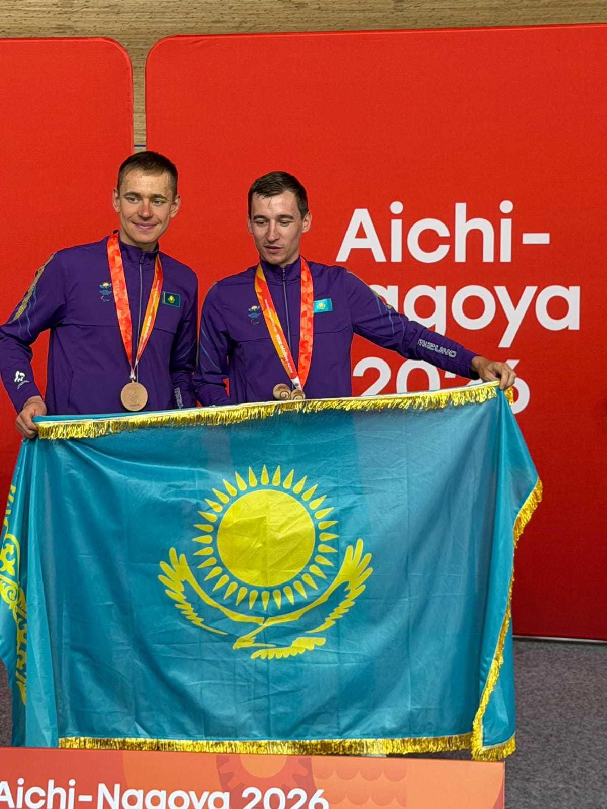 Қазақстан туын ұстаған, медаль алған екі спортшы. Фонда «Aichi-Nagoya 2026» жазуы бар.