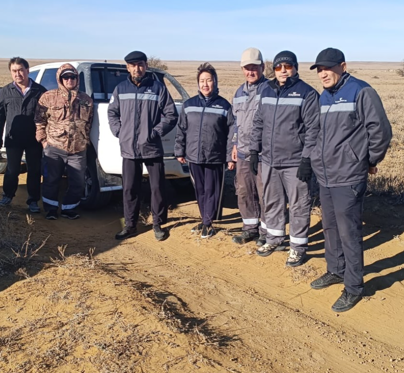 Ақ жол жиегінде, ақ түсті көліктің жанында тұрған жұмыс киіміндегі бір топ адам.