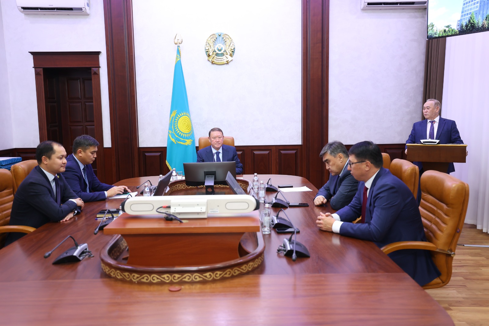 Қазақстан Республикасының мемлекеттік рәміздері бар залда өткізіліп жатқан ресми жиналыс.