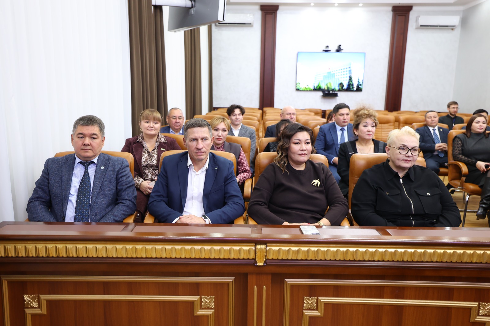 Жиналыс залында отырған ресми киінген адамдар тобы.