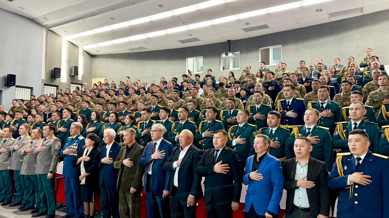 Группа военных в форме и гражданских лиц стоят в зале, приложив правую руку к сердцу.