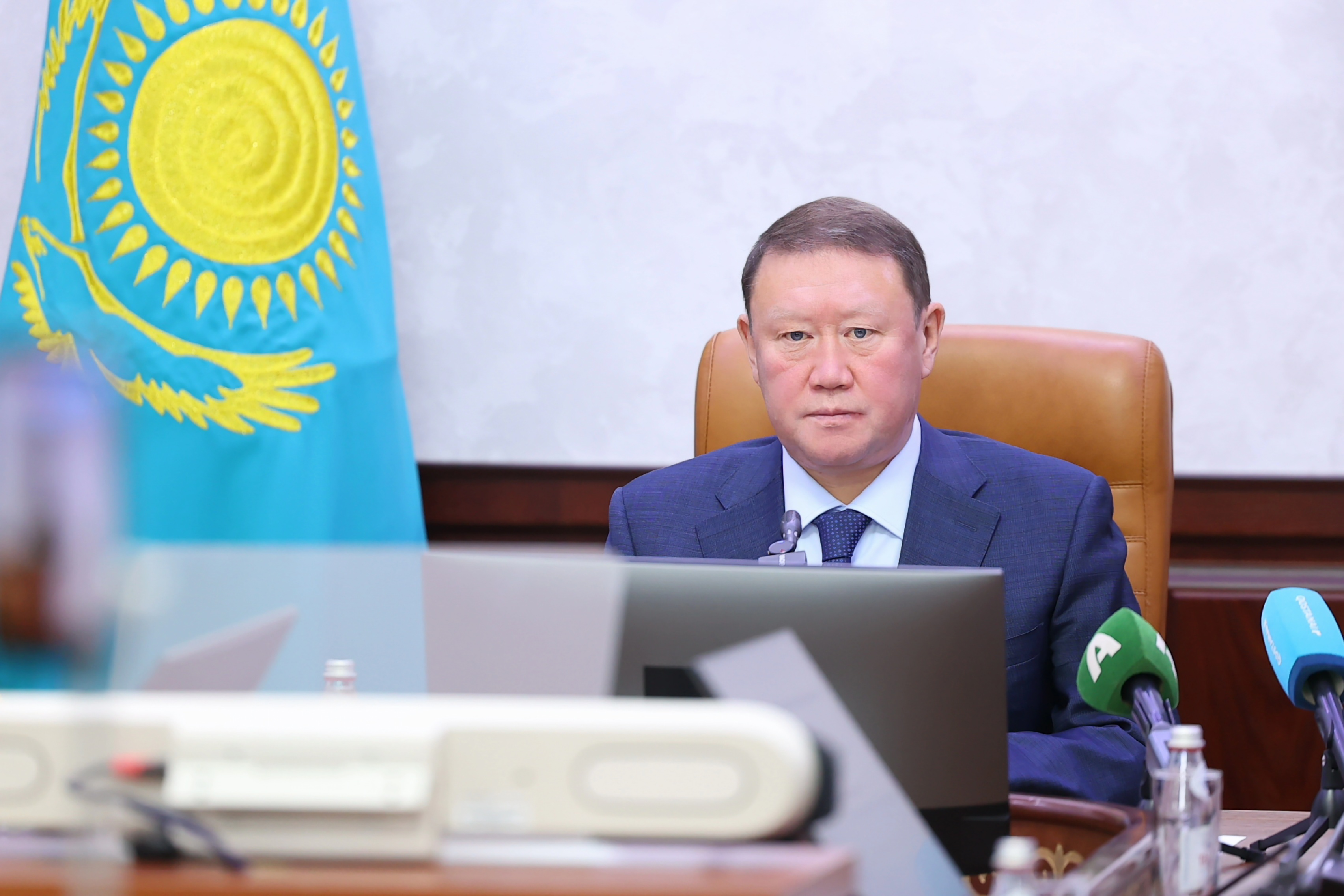 Қазақстан Республикасының туының жанында ноутбук алдында отырған костюм киген ер адам.