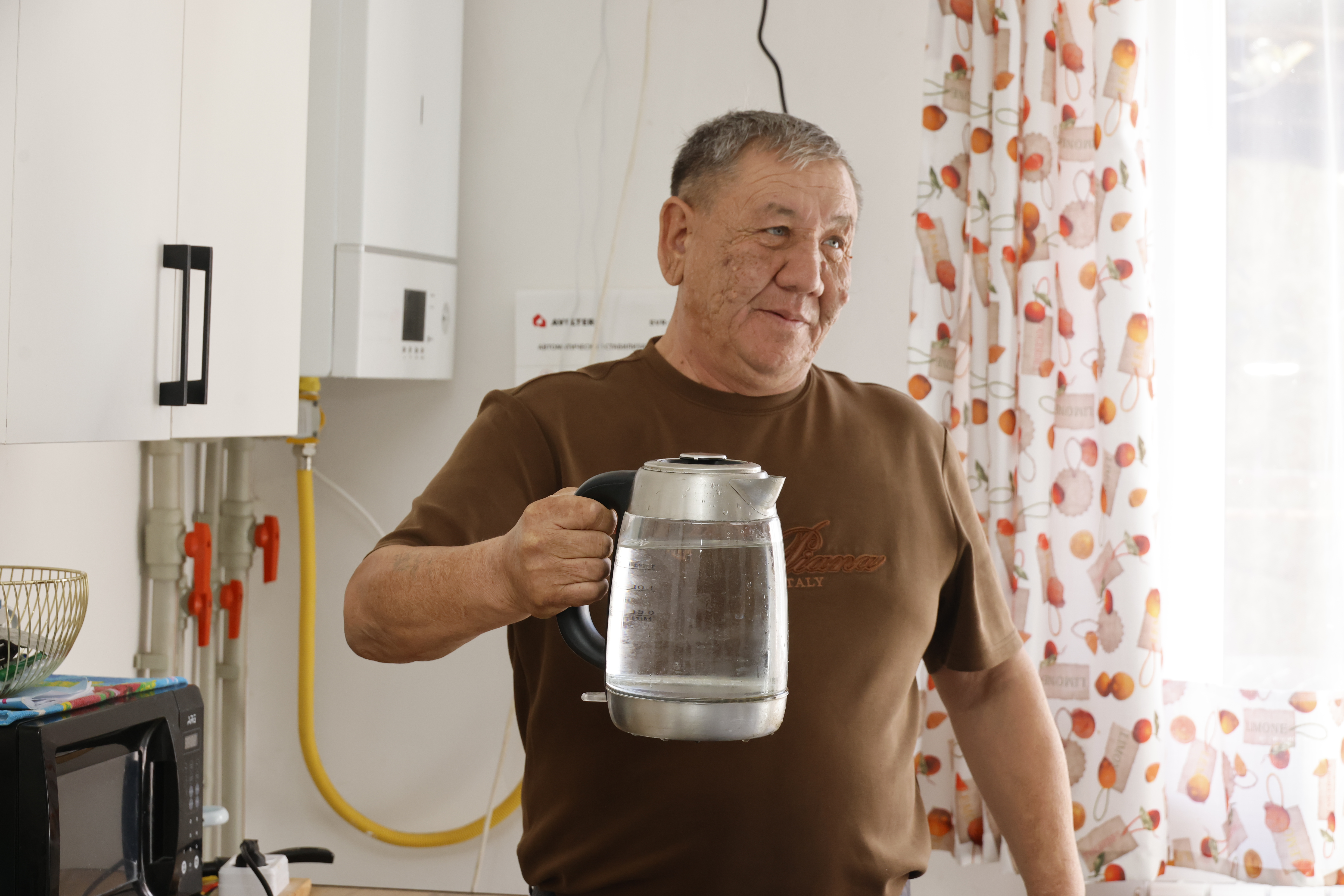 Пожилой мужчина в коричневой футболке держит в руке стеклянный электрический чайник на кухне.