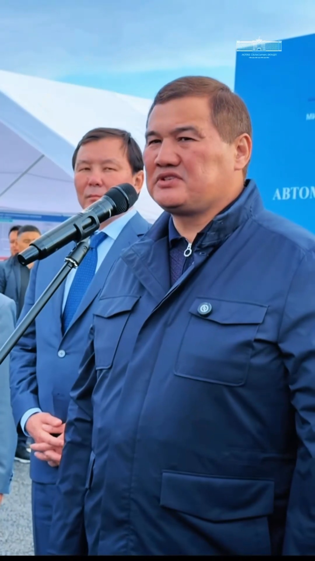 Микрофон алдында сөйлеп тұрған көк куртка киген ер адам.