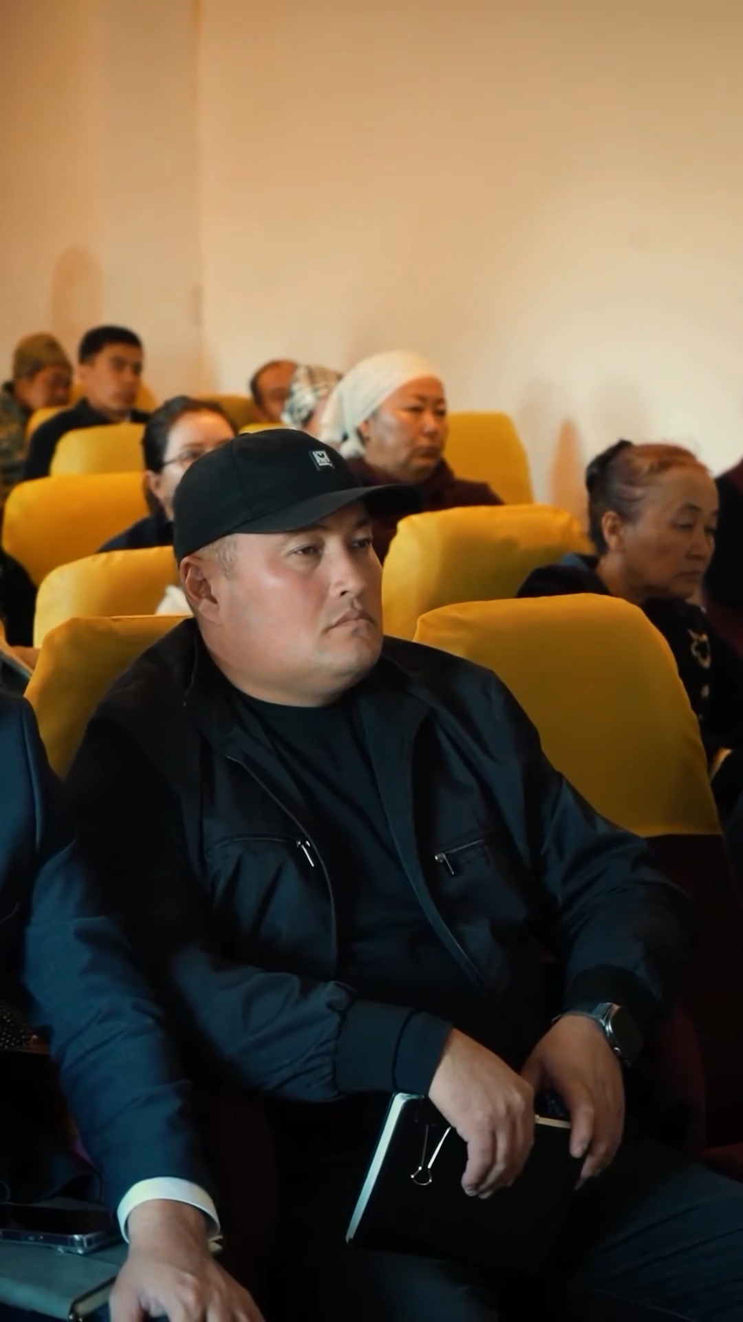 Қара кепкен және қара кепка киген ер адамның аудиторияда отырған сәті.