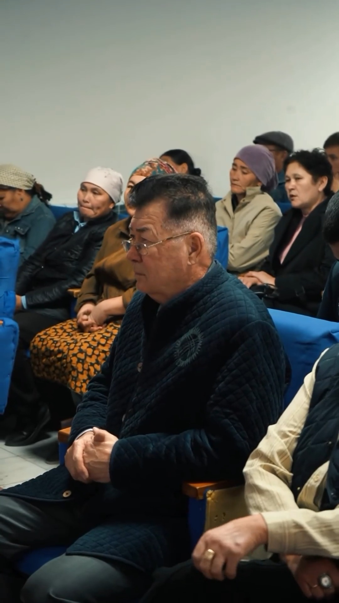 Көк орындықтарда отырған адамдар тобы, алдыңғы планда қою түсті күрте киген ер адам.