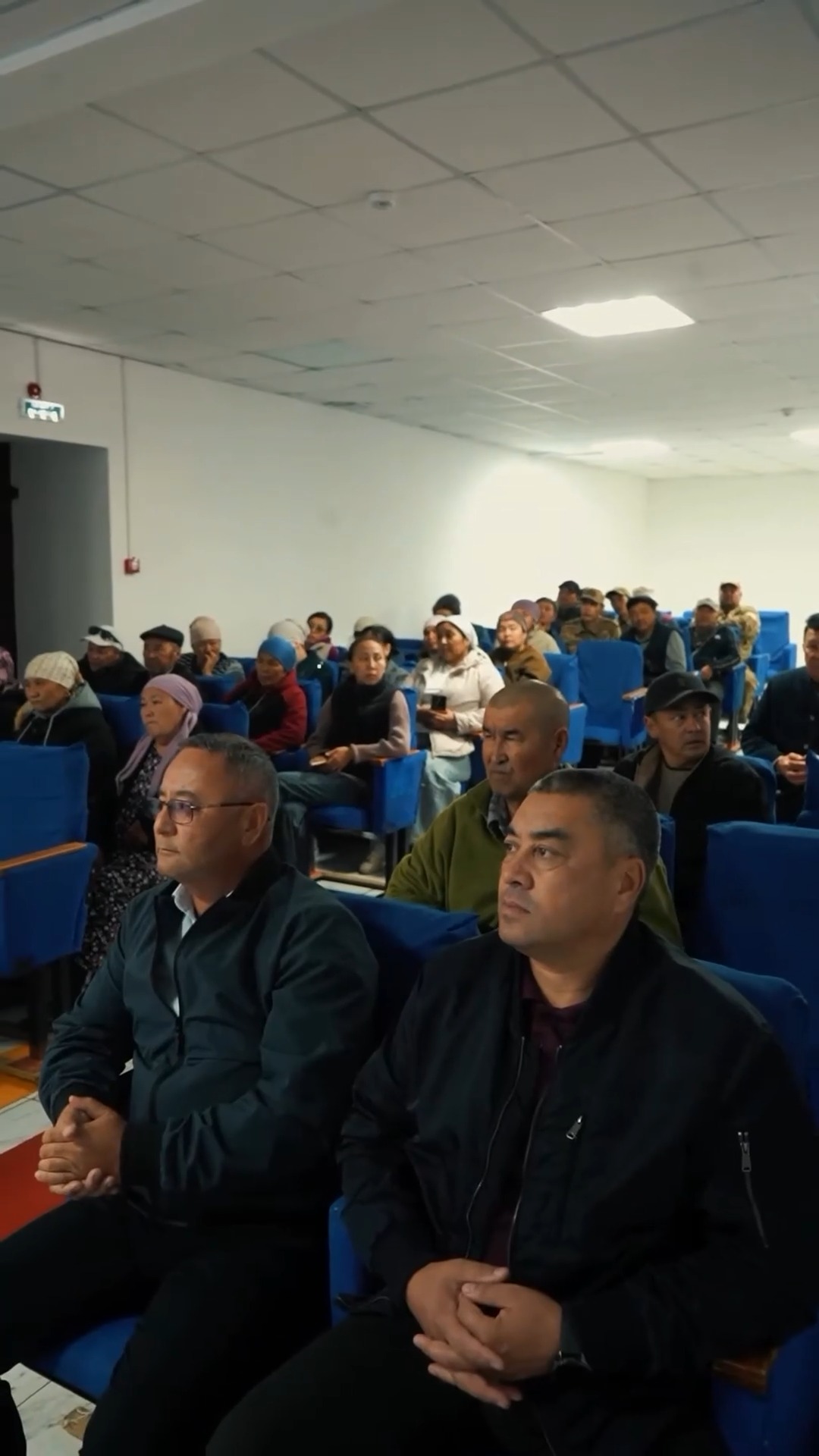 Жиналыс залындағы көк орындықтарда отырған адамдар тобы.