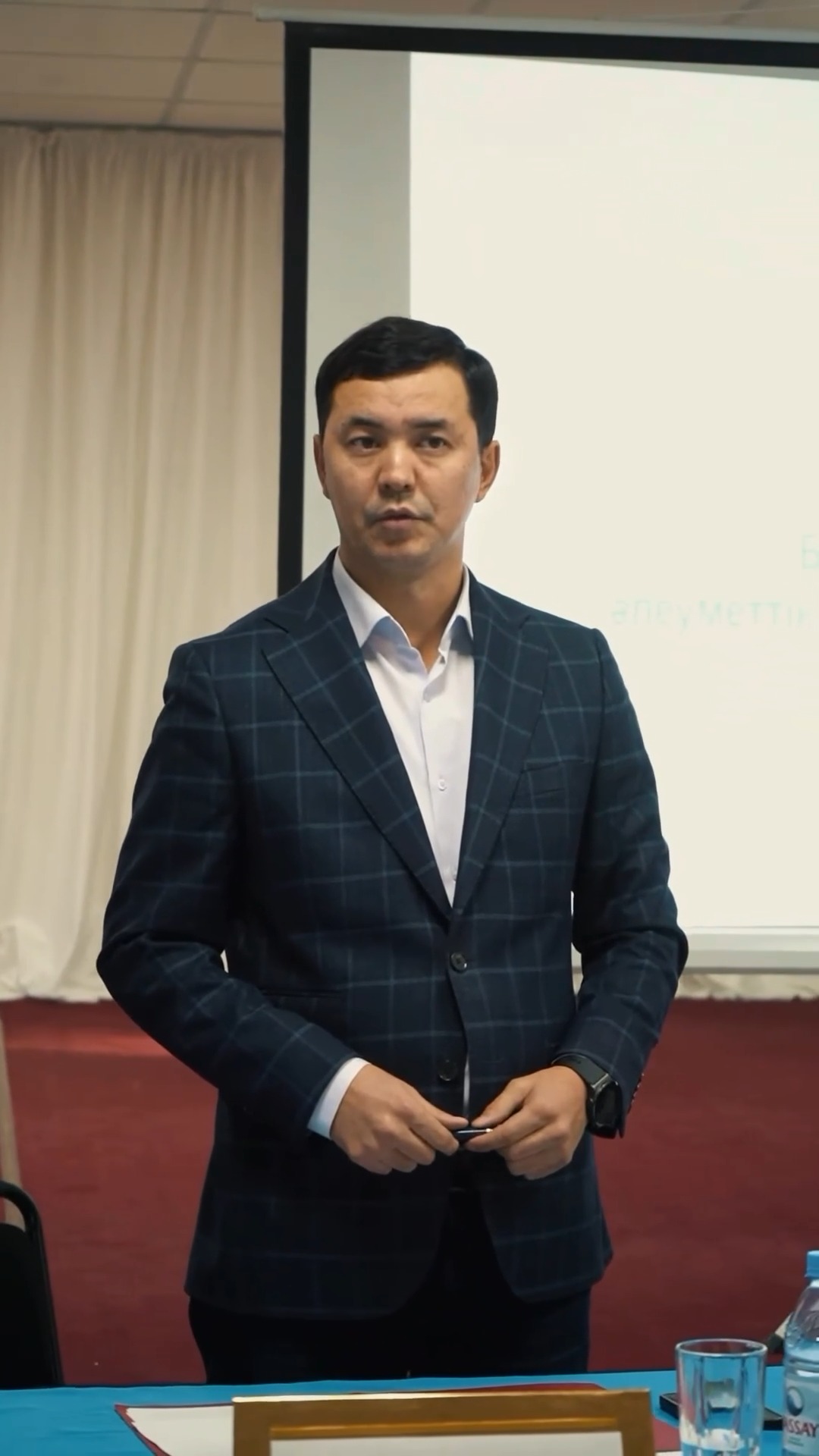 Көк торлы костюм киген ер адам презентация алдында тұр.