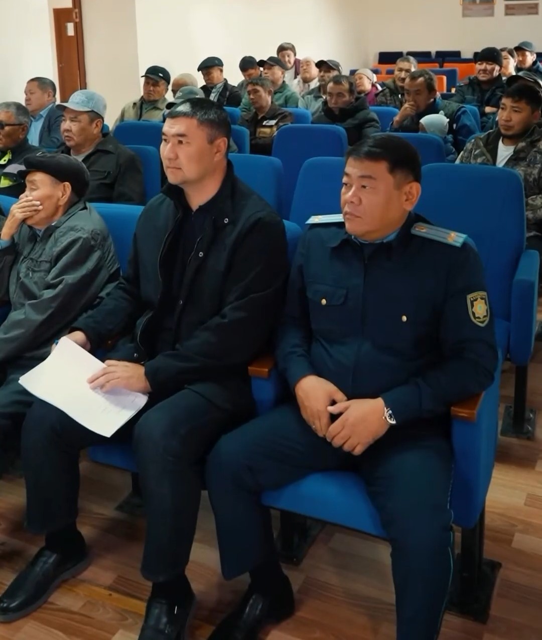 Көк орындықтарда отырған полиция қызметкері мен қара киінген ер адам және жиналысқа қатысушылар.