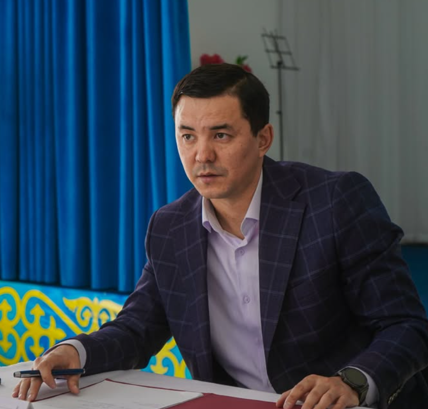 Көк перде алдында үстел басында отырған, қолына қалам ұстаған костюм киген ер адам.