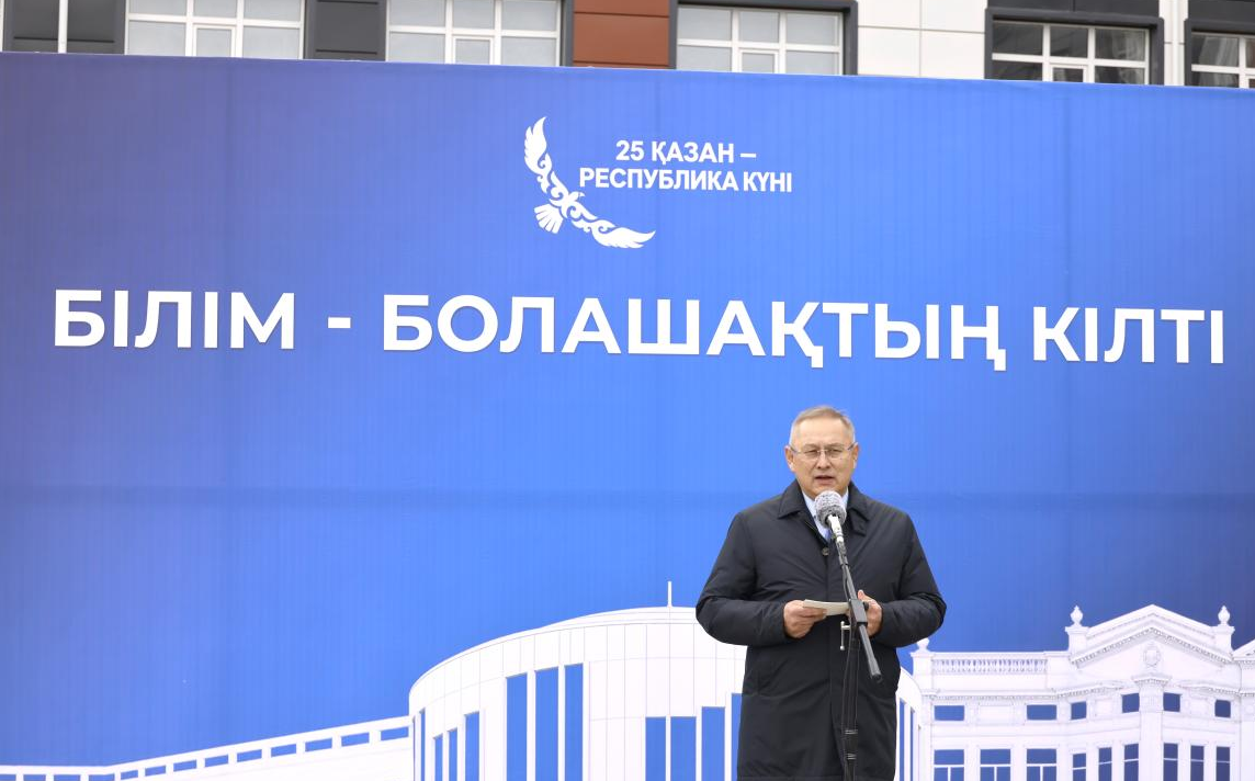 Көк түсті «Білім - болашақтың кілті» деген жазуы бар баннер алдында сөйлеп тұрған ер адам.