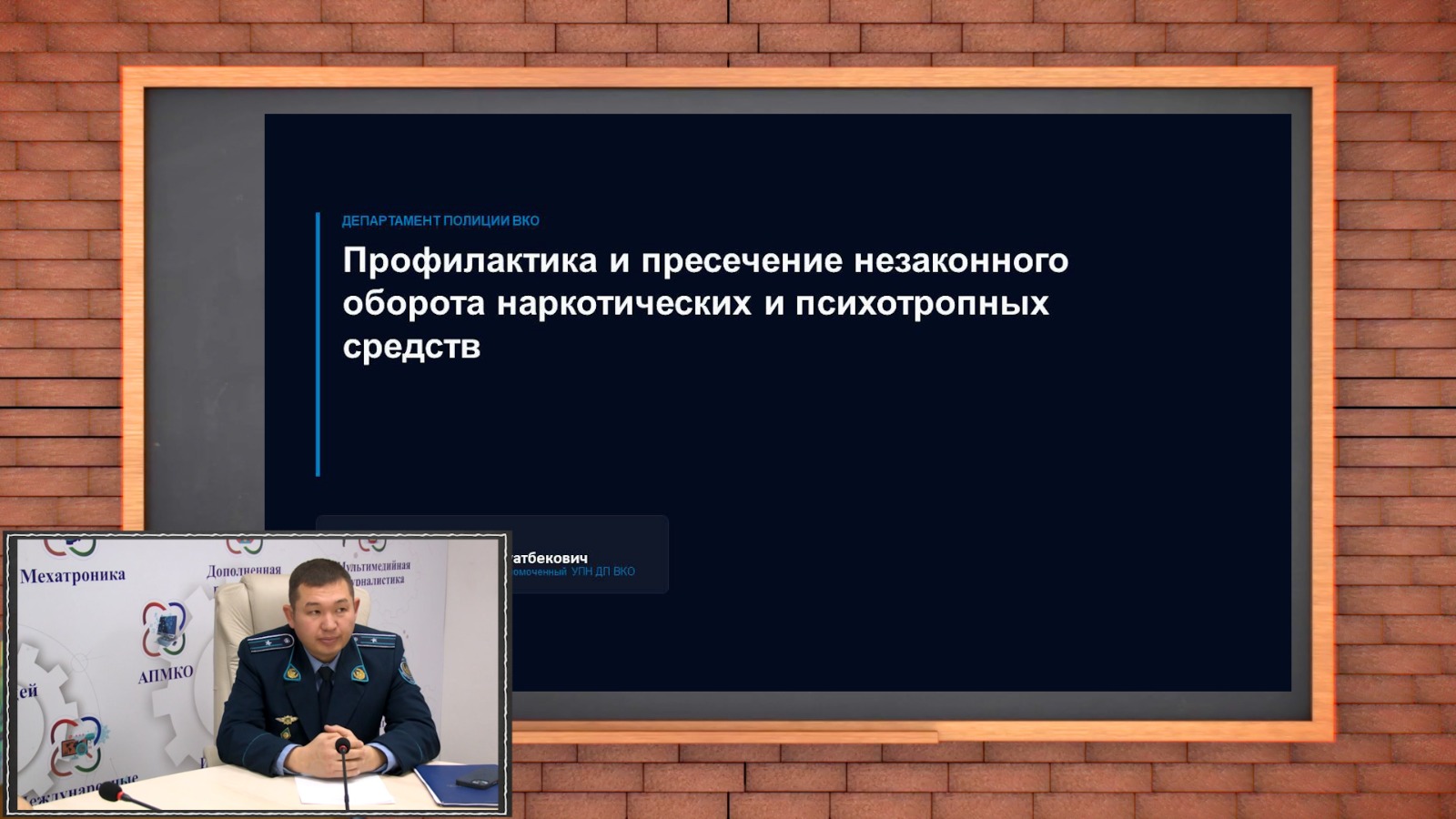 Есік қолындағы презентация алдында отырған полиция қызметкері. Тақырыбы: «Есірткі және психотроптық заттардың заңсыз айналымын алдын алу».