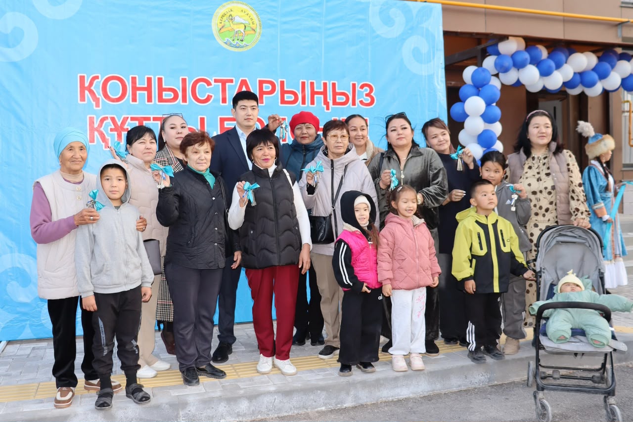 Жаңа пәтерлерді тапсыру рәсіміндегі адамдар тобы. Артында «Қоныстарыңыз құтты болсын!» деген жазуы бар көк баннер тұр.