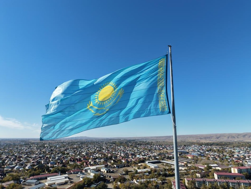 Көк аспан астында, қала көрінісінің фонында желбіреген Қазақстан Республикасының мемлекеттік туы.