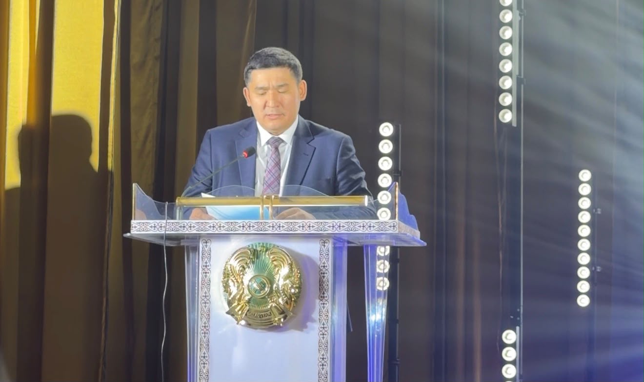 Көк костюм киген ер адам трибунада сөйлеп тұр.