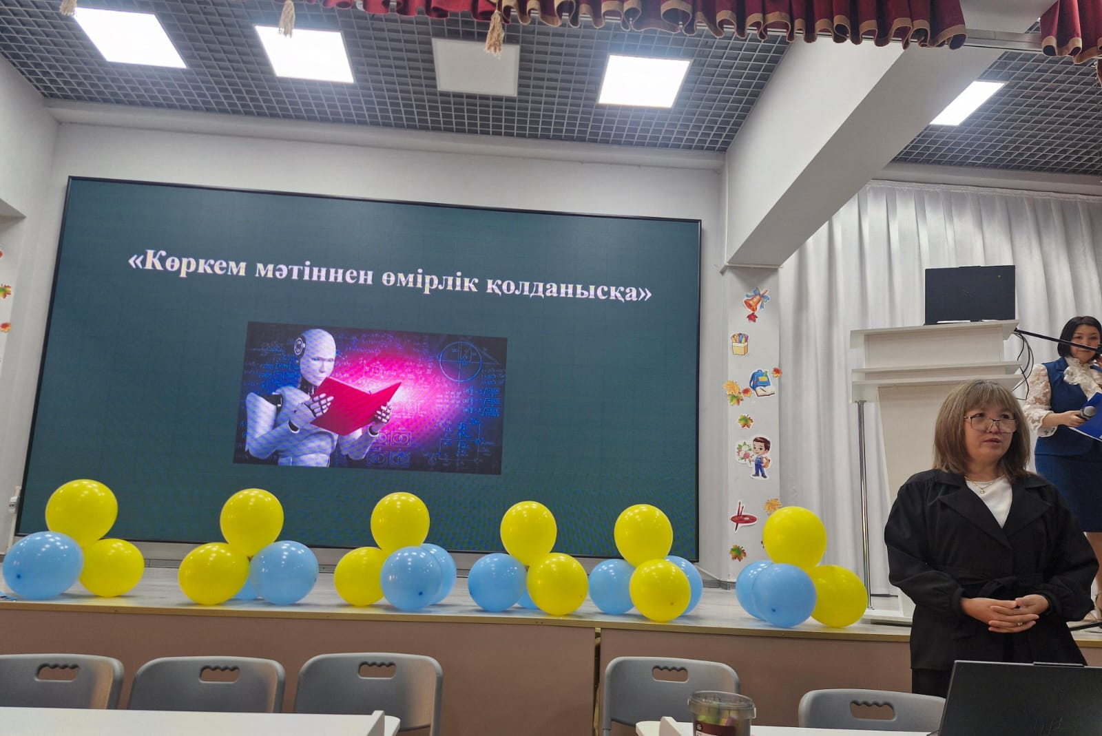 Экранда «Көркем мәтіннен өмірлік қолданысқа» тақырыбындағы презентация және алдында тұрған әйел адам.