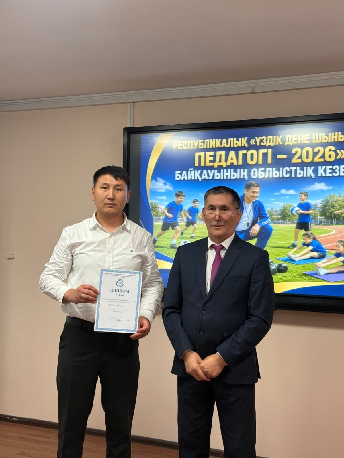 Диплом алған жас маман мен костюм киген ер адамның «Үздік дене шынықтыру педагогі – 2026» жазуы бар экран алдындағы суреті.