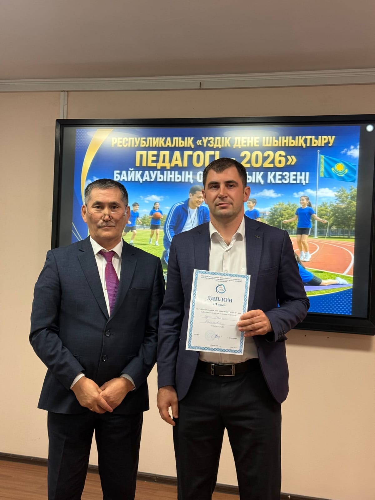 Костюм киген екі ер адам. Біреуі қолына «Үздік дене шынықтыру педагогі» байқауының дипломын ұстап тұр.