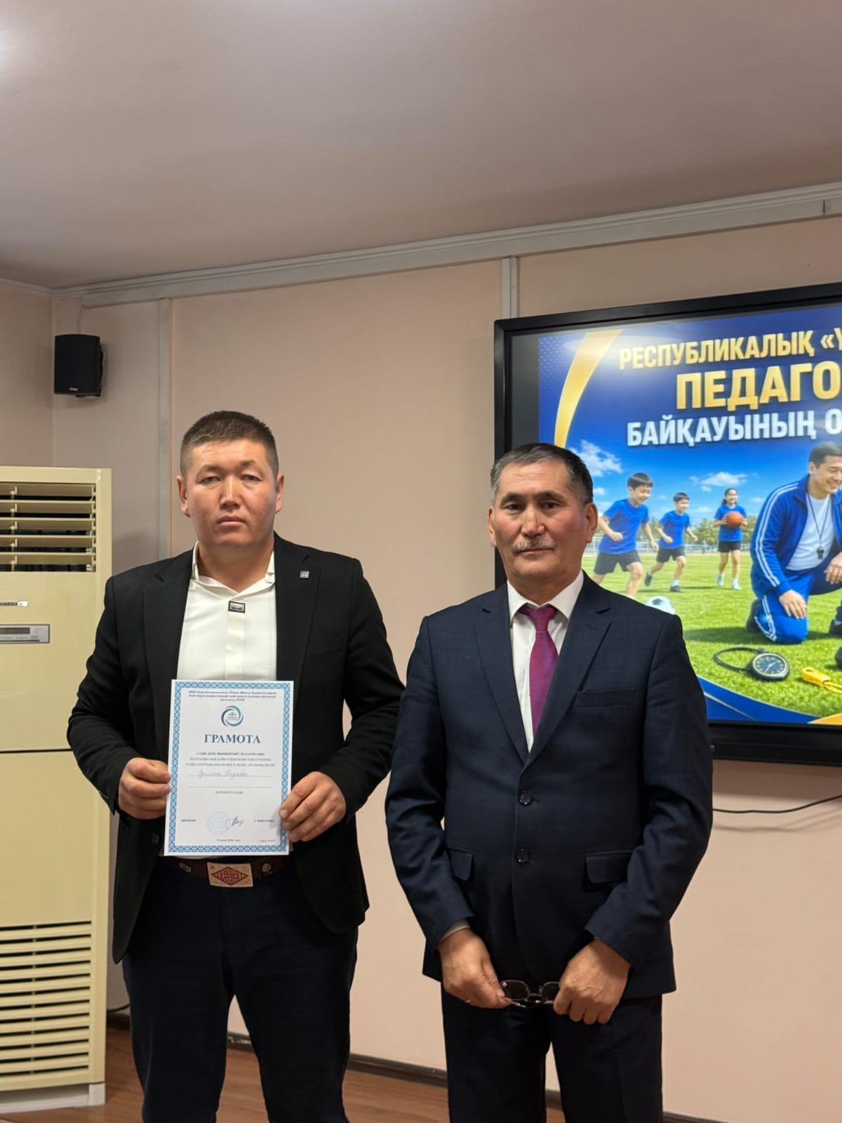 Костюм киген екі ер адам, бірі қолына грамота ұстап тұр. Артында презентация экраны бар.