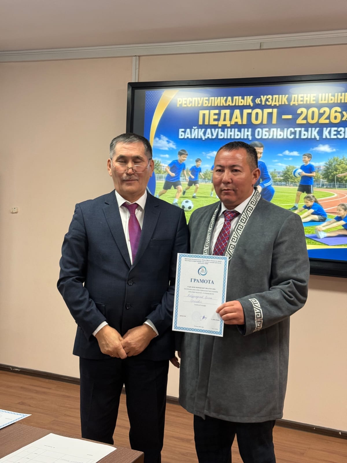 Костюм киген екі ер адам: бірі грамота ұстап тұр, артқы жақта «Үздік дене шынықтыру педагогі – 2026» жазуы бар экран.