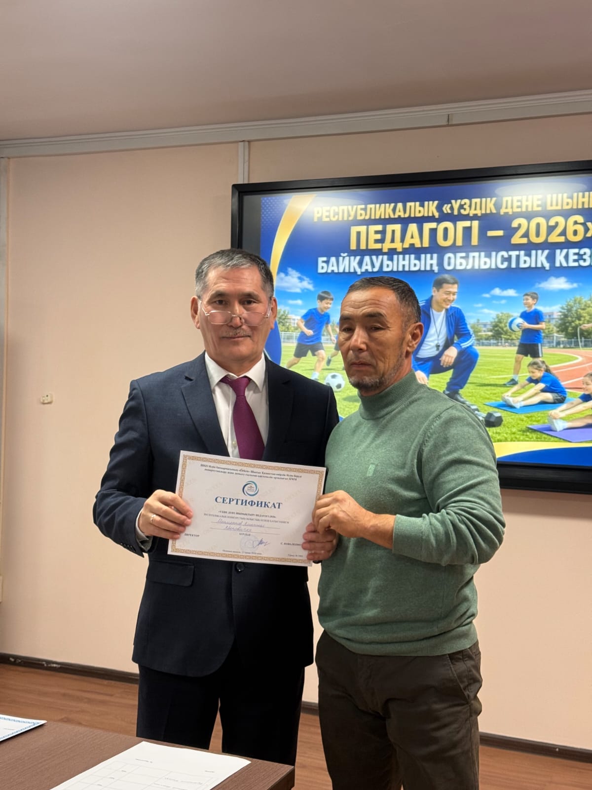 Екі ер адамның «Үздік дене шынықтыру педагогі – 2026» байқауында сертификатты табыстасу сәті.