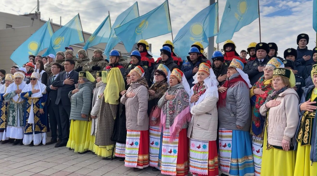 Қазақстан туларының алдында қолы жүрегінде тұрған ұлттық киімді әйелдер мен ресми киімдегі ерлер тобы.
