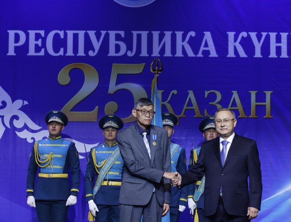 Республика күніне арналған салтанатты жиында екі ер адамның қол алысуы. Фонда «25 ҚАЗАН» жазуы бар.