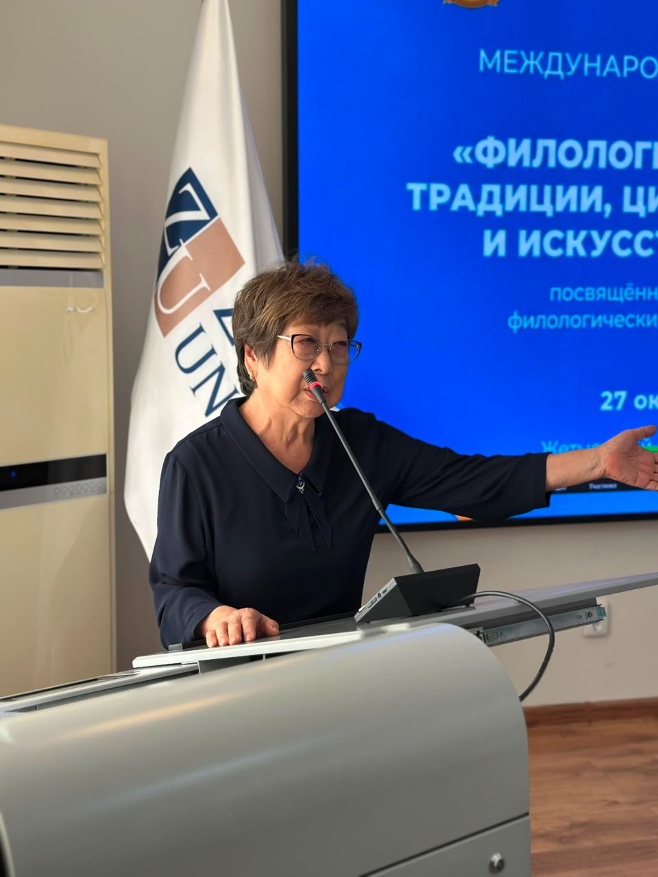 Трибунада сөйлеп тұрған әйел адам. Артында презентация экраны мен университет туы.