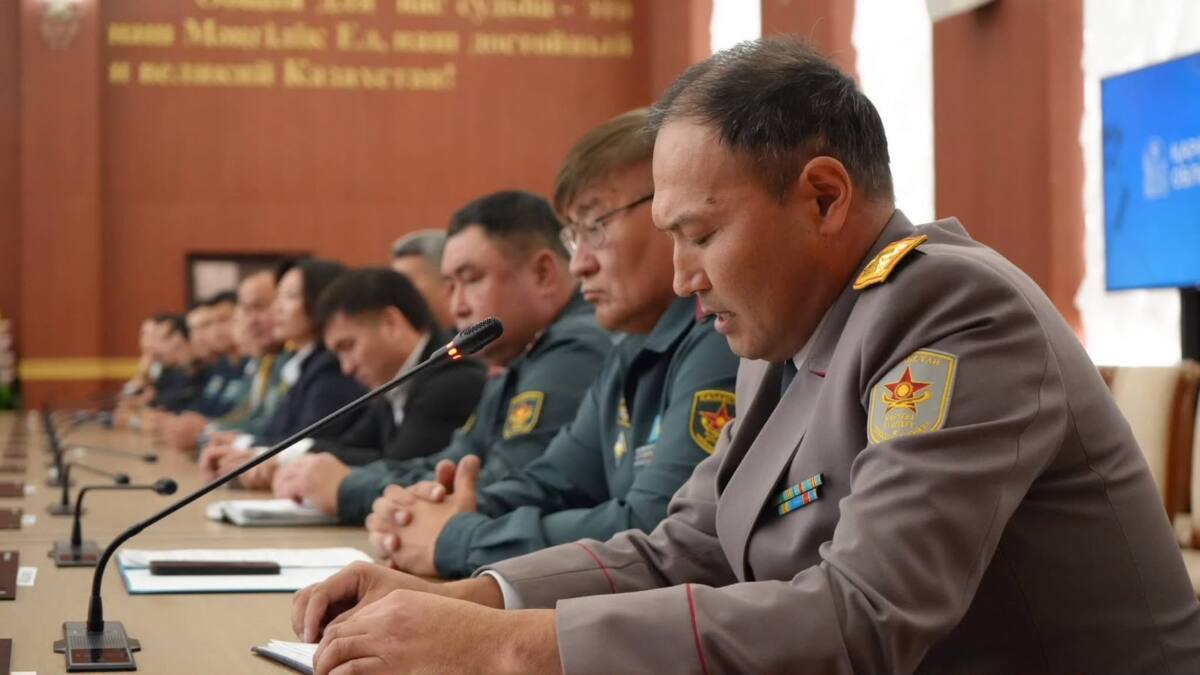 Жиналыста сөйлеп жатқан әскери формадағы ер адам және оның қасында отырған әріптестері.