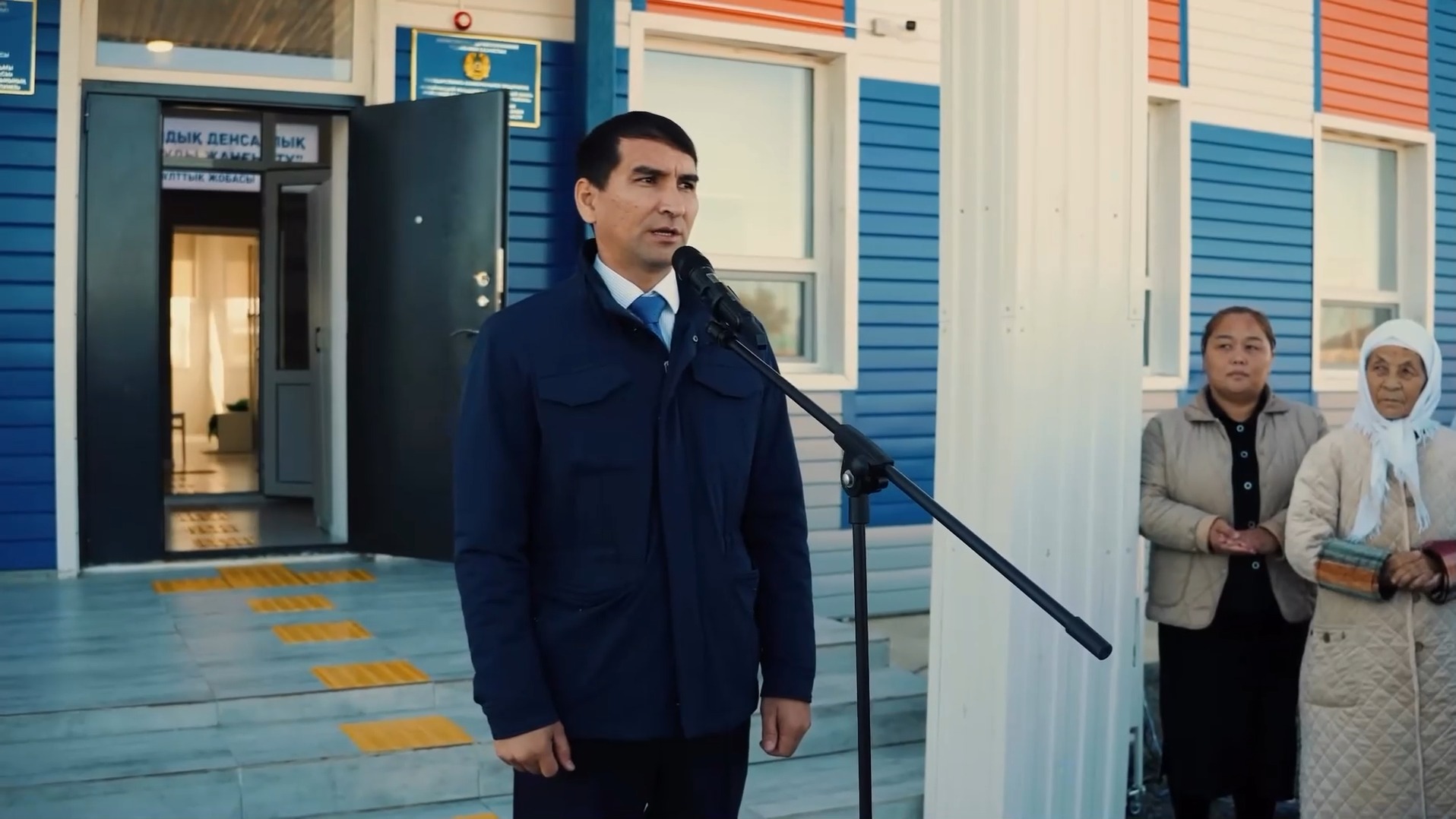 Микрофон алдында сөйлеп тұрған ер адам және оның артында тұрған екі әйел.