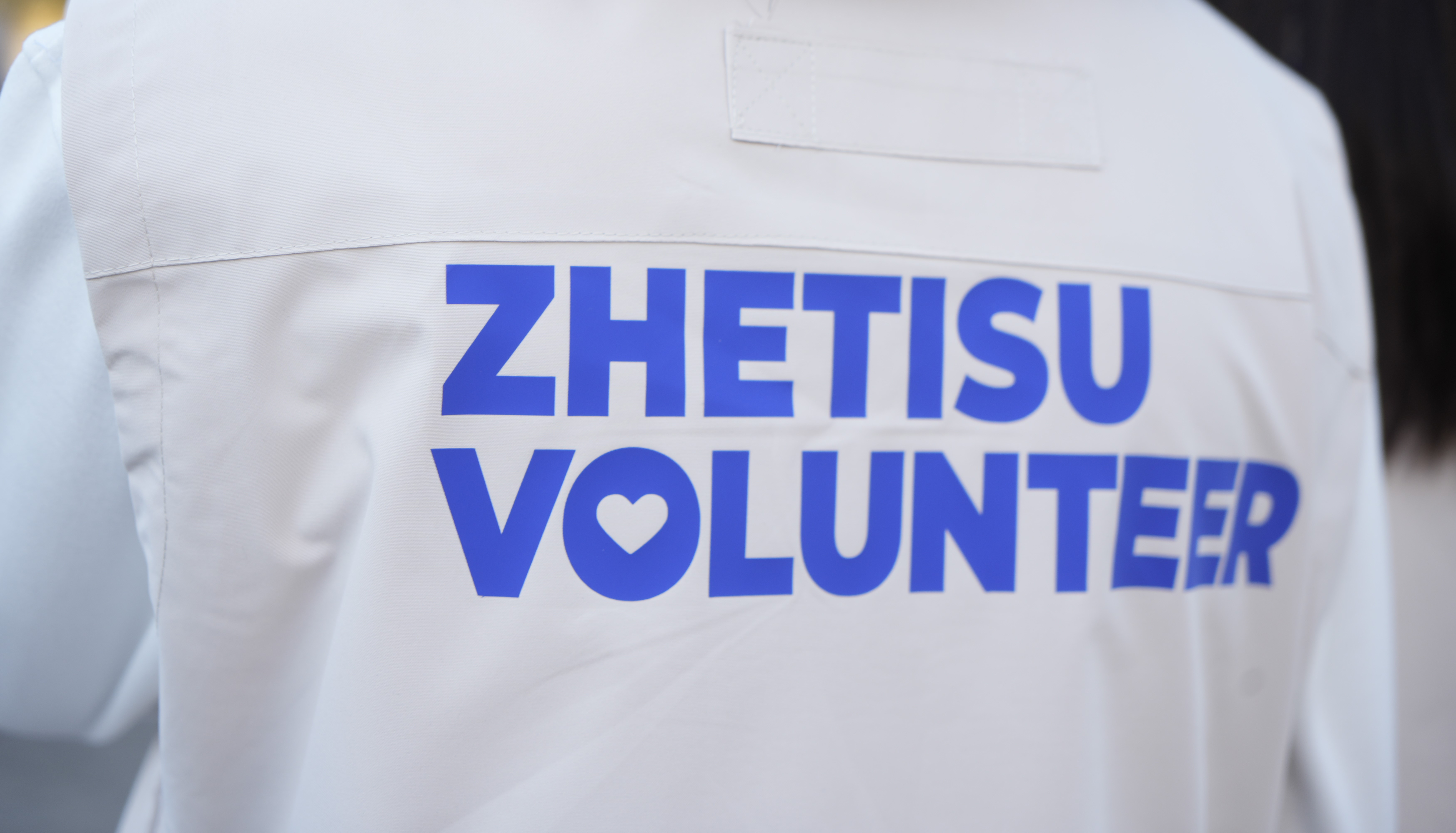 Спина человека в белой жилетке с синей надписью «ZHETISU VOLUNTEER».