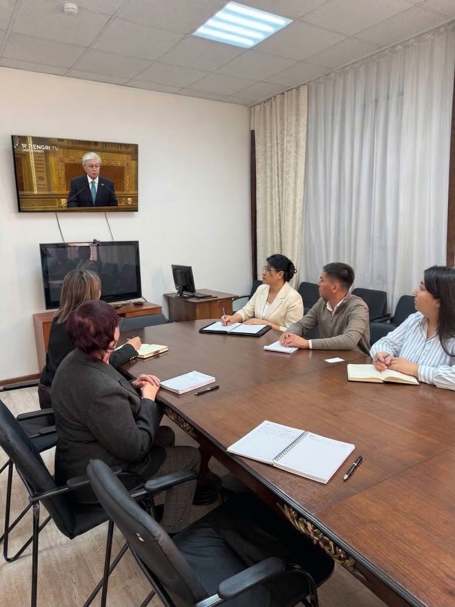 Кеңседегі үстел басында отырған адамдардың экрандағы спикерді тыңдап, жазбалар жасап жатқан сәті.