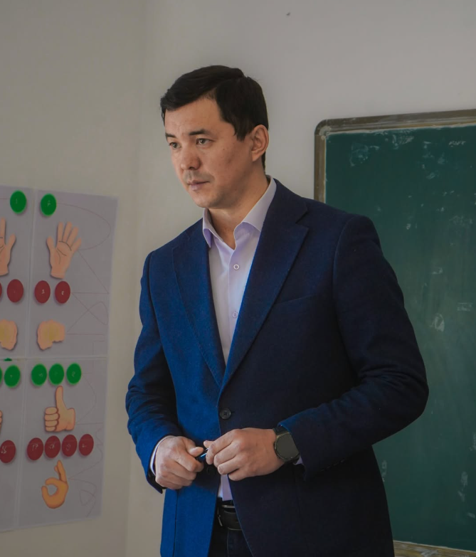 Көк костюм киген ер адам сыныпта тақтаның жанында тұр.