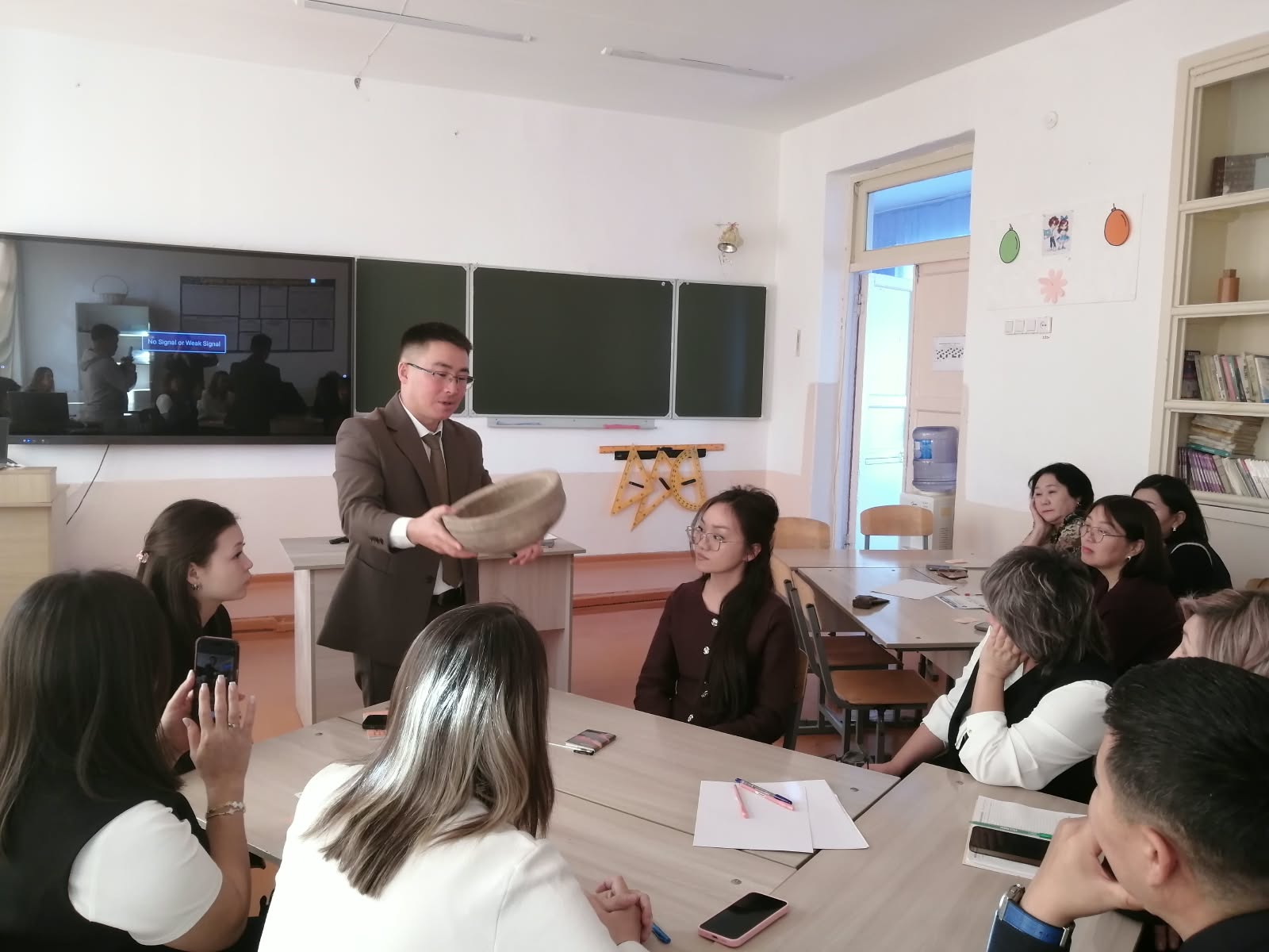Сыныпта костюм киген мұғалім оқушыларға ұлттық бас киімді көрсетіп, сабақ жүргізіп жатыр.