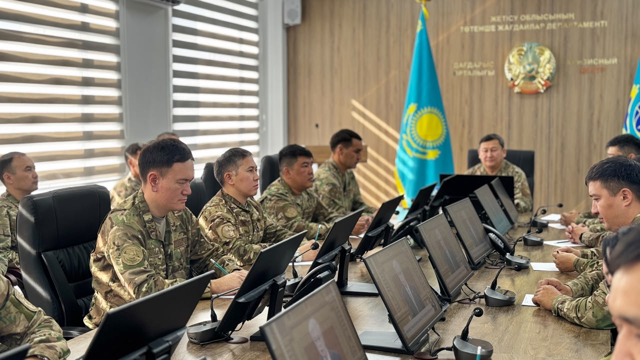 Группа военных в камуфляже за столом с мониторами в кабинете на фоне флага Казахстана.