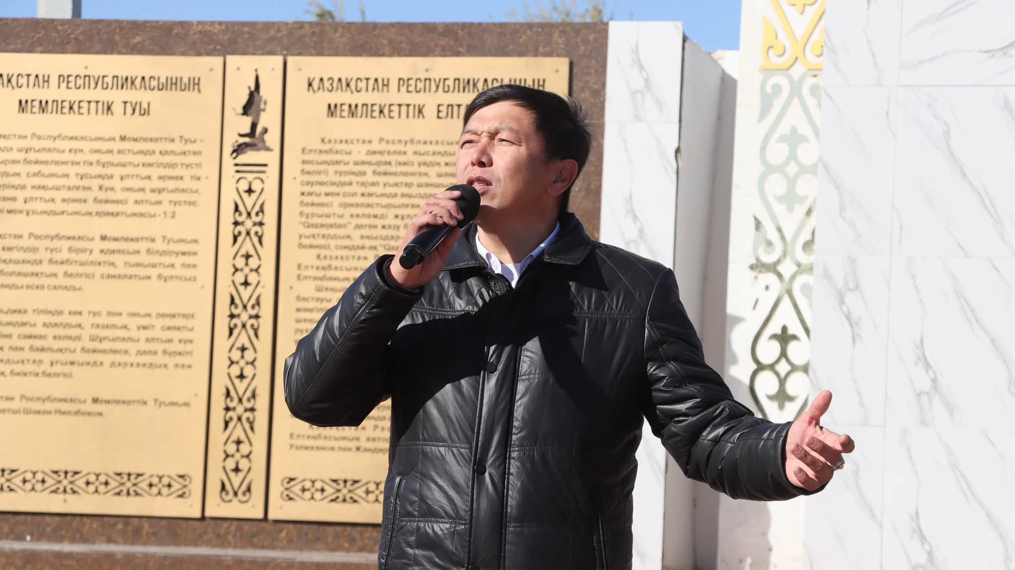 Ер адам микрофонмен ән айтып  тұр, артында Қазақстан Республикасының мемлекеттік рәміздері жазылған тақталар.