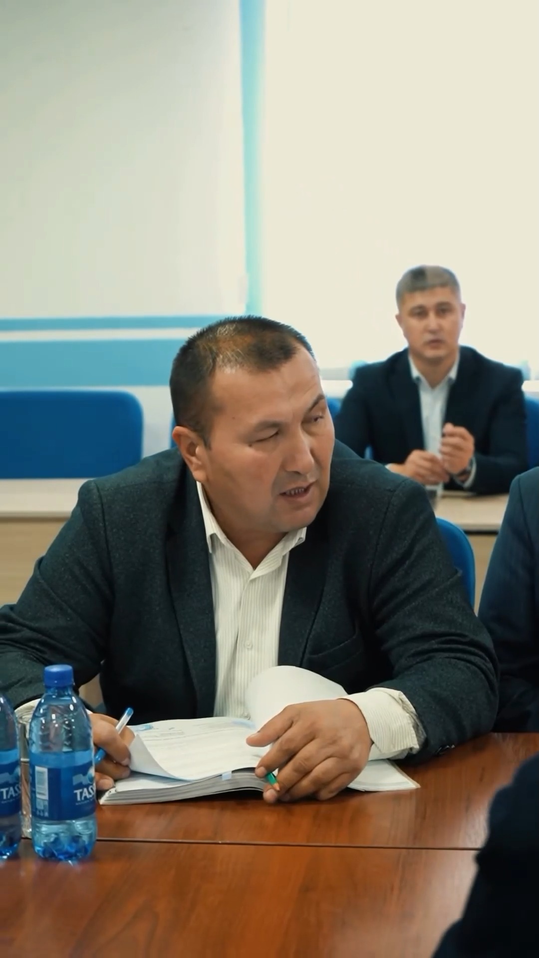 Жиналыс кезінде сөйлеп жатқан костюм киген ер адам.