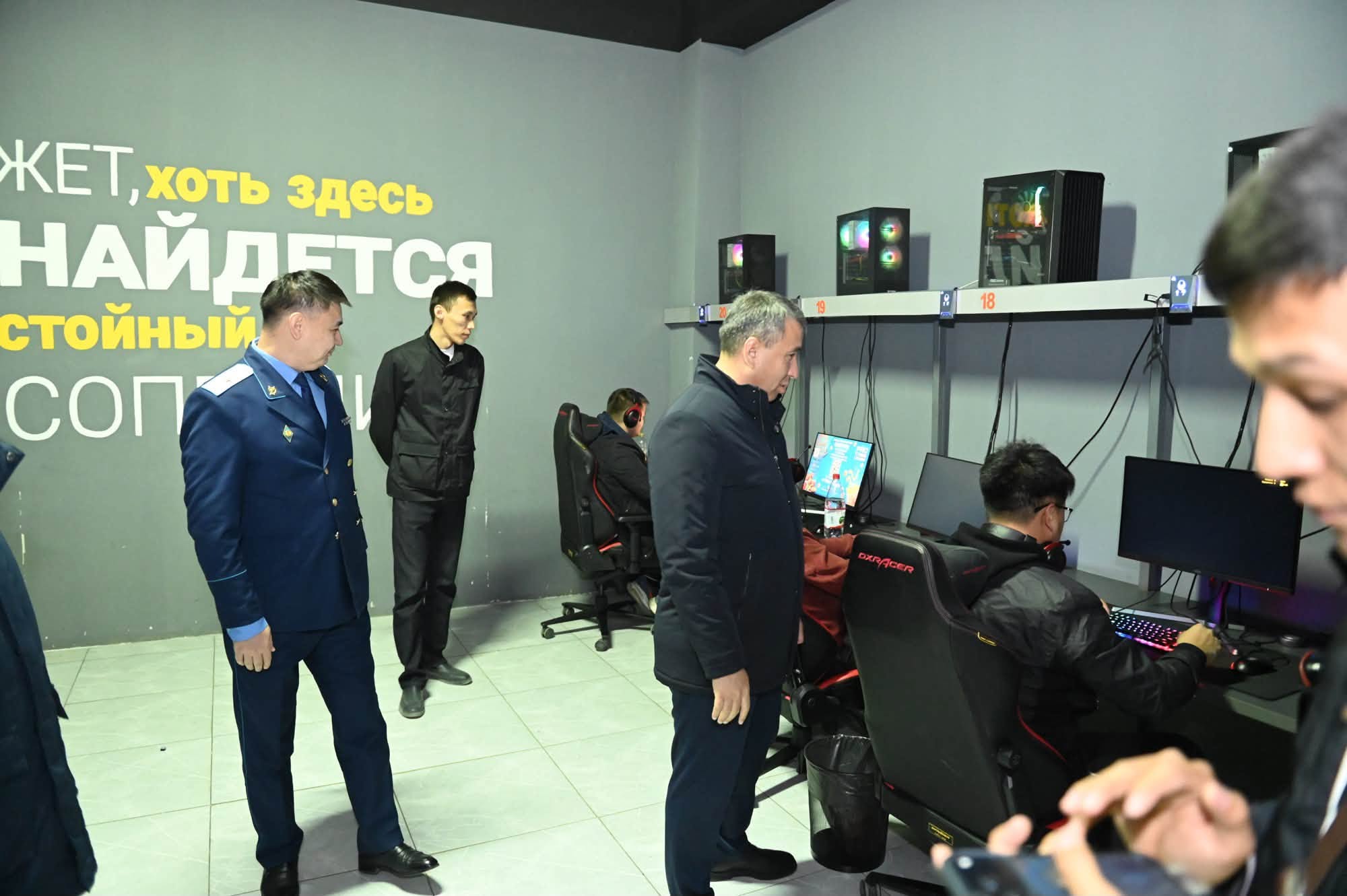 Киберспорттық клубда компьютер алдында отырған ойыншылар мен оларды бақылап тұрған ресми тұлғалар.