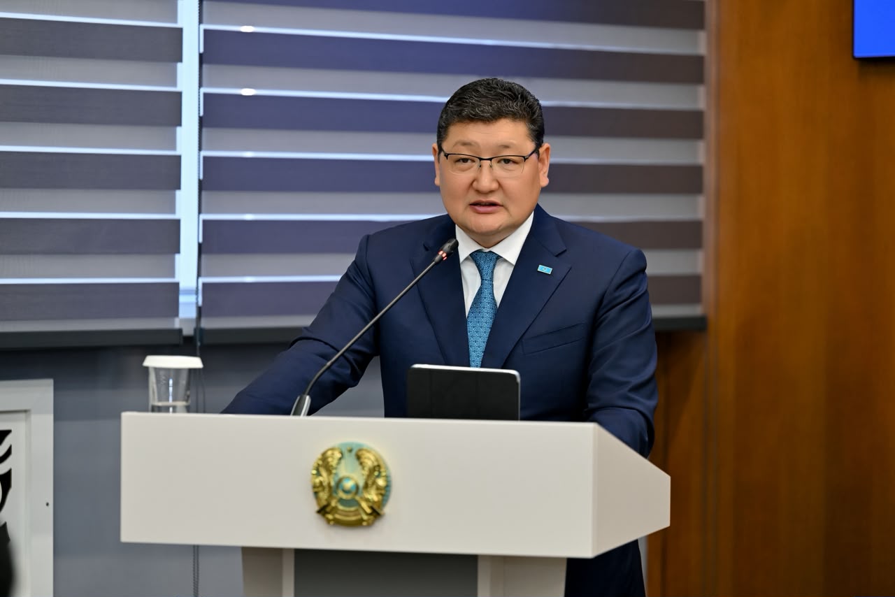 Көзілдірік пен костюм киген ер адам трибунада сөйлеп тұр.