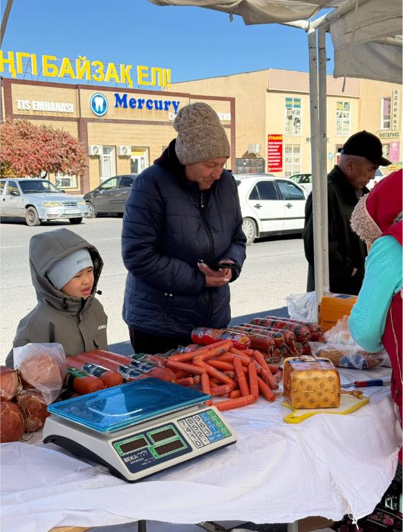 Көшедегі базарда шұжықтар мен колбасалар сатылып жатыр, сатып алушылар тұр.