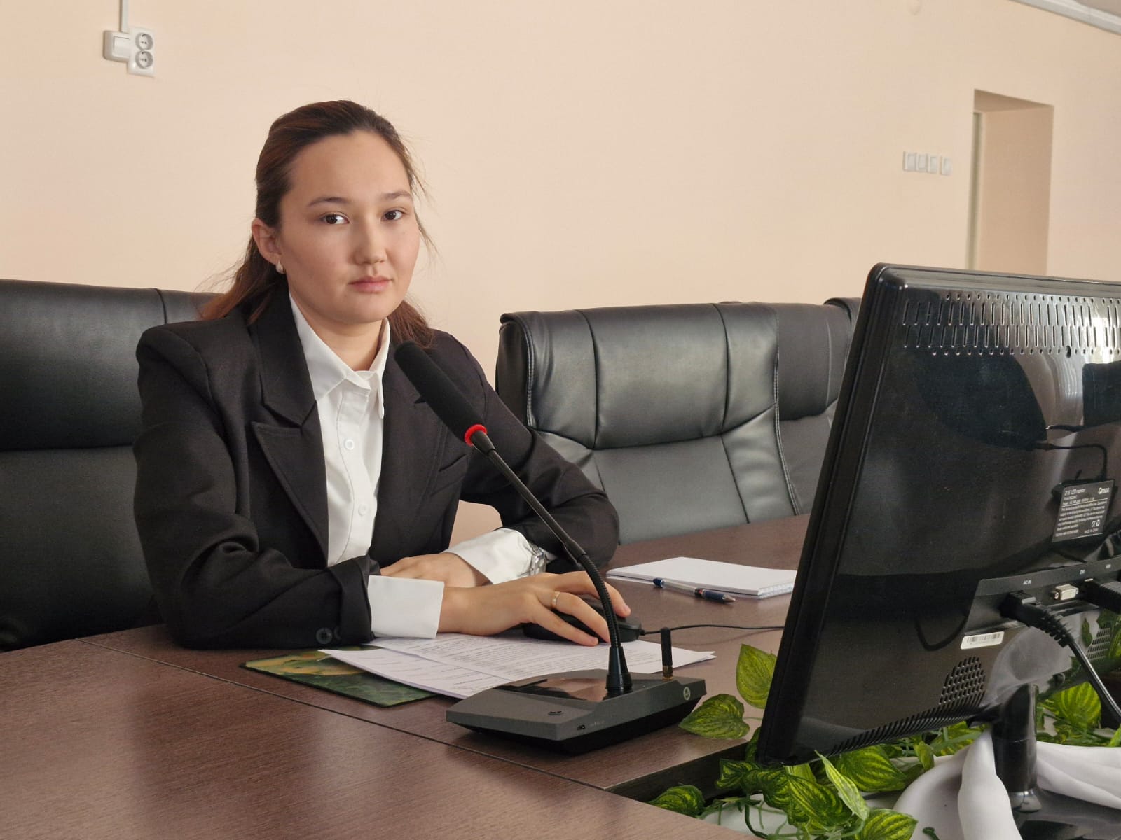 Кеңседе компьютер мен микрофон алдында отырған ресми киінген әйел.