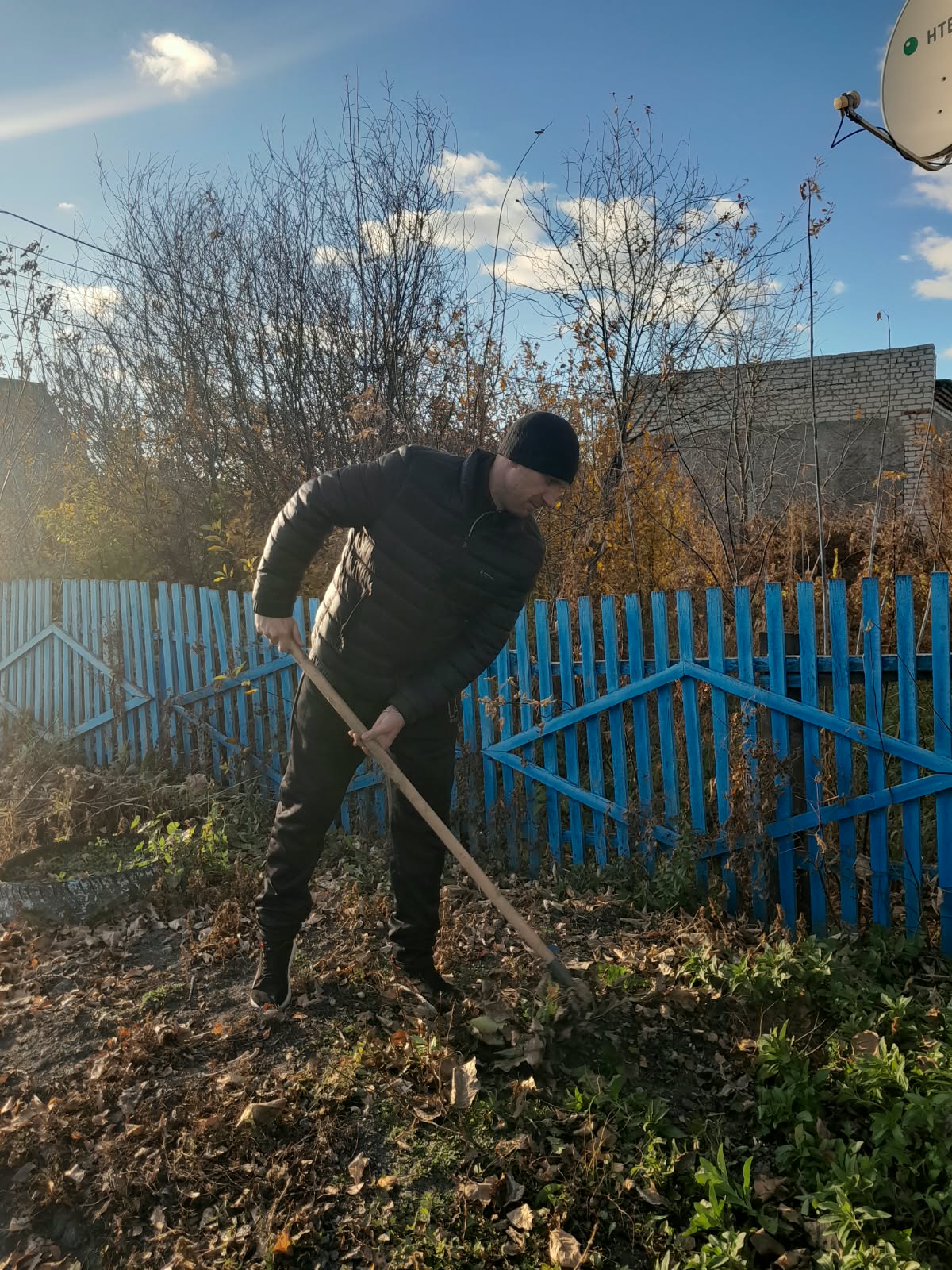 Қара күрте мен қалпақ киген адам көк қоршаудың фонында тырмамен жұмыс істейді.