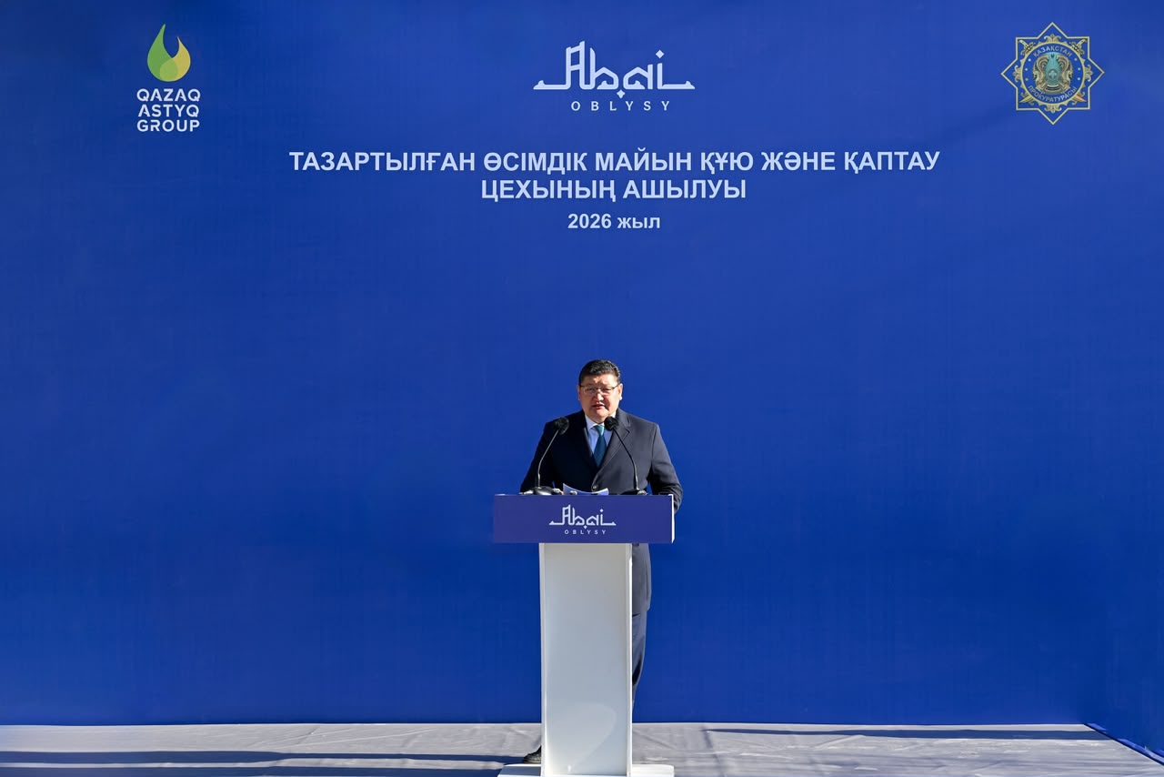 Көк фонды сахнада трибунада сөйлеп тұрған ер адам. Артында «Тазартылған өсімдік майын құю және қаптау цехының ашылуы» жазуы бар.