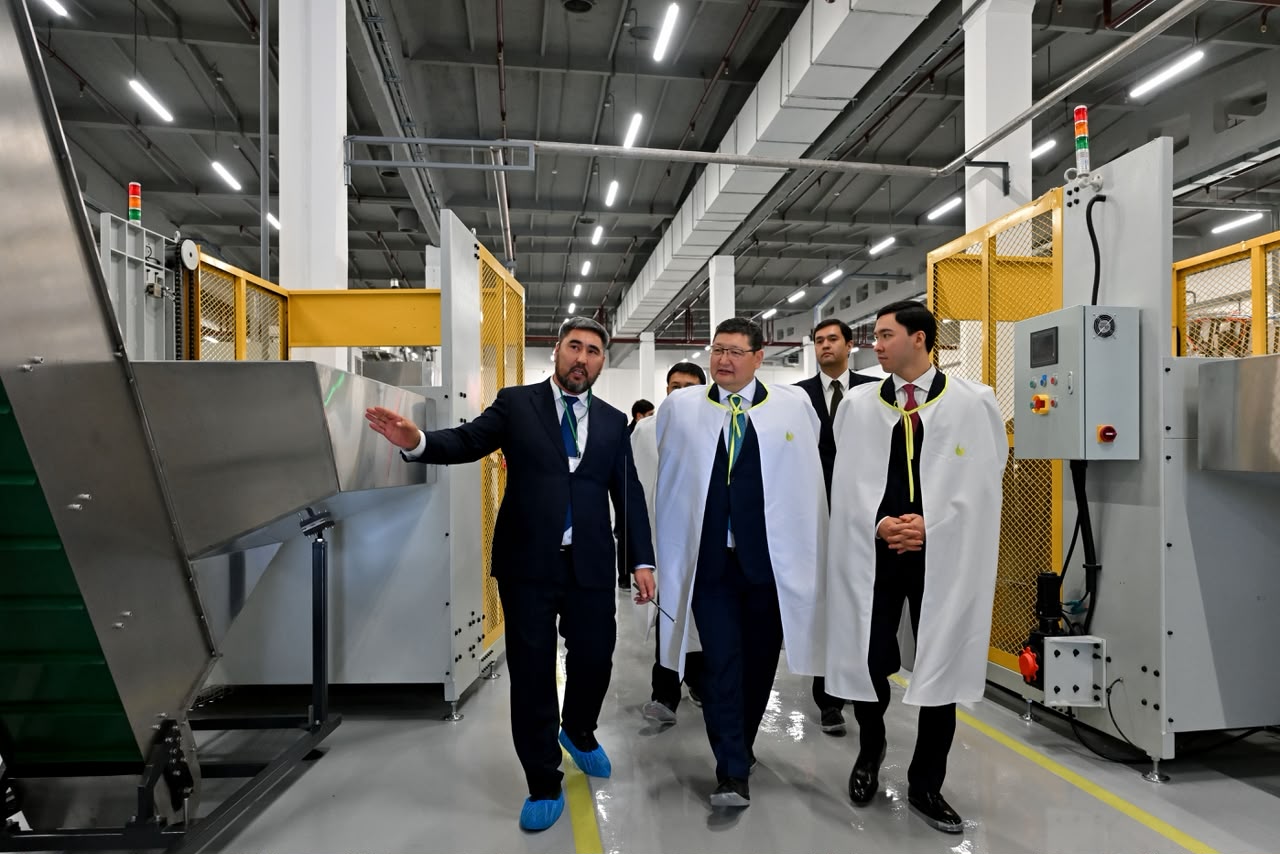 Зауыт цехіндегі өндірістік жабдықтарды аралап жүрген костюм киген еркектер мен академиялық киімдегі мамандар.