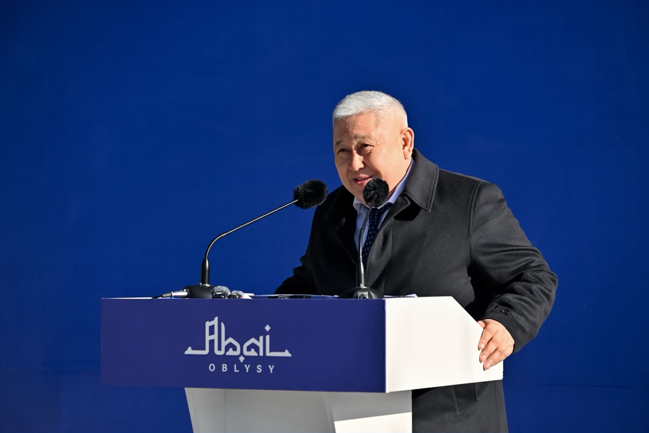 Көк фон алдындағы трибунада сөйлеп тұрған қара пальто киген ер адам.