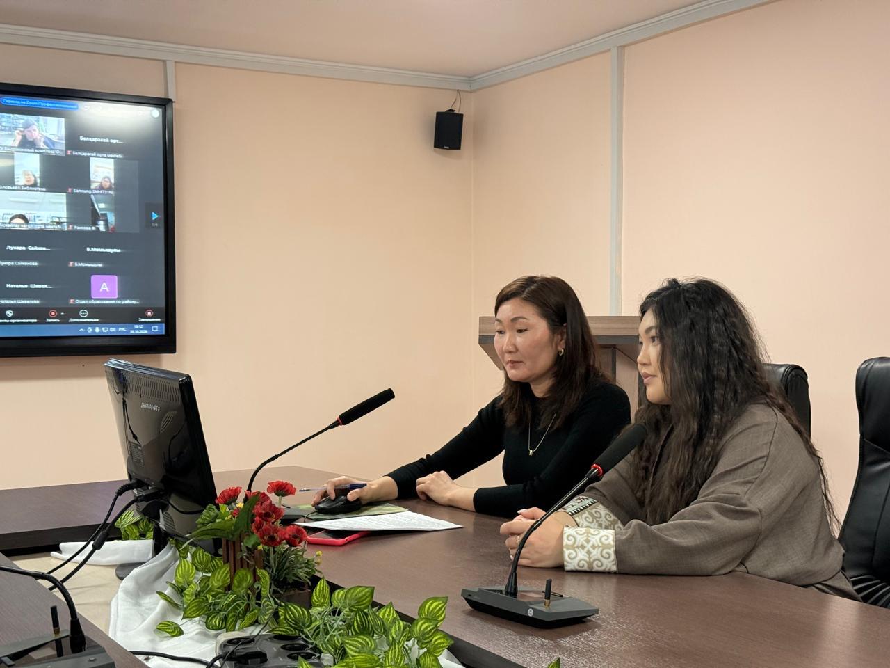 Екі әйел онлайн мәжіліске қатысып, монитор мен экранға қарап отыр.