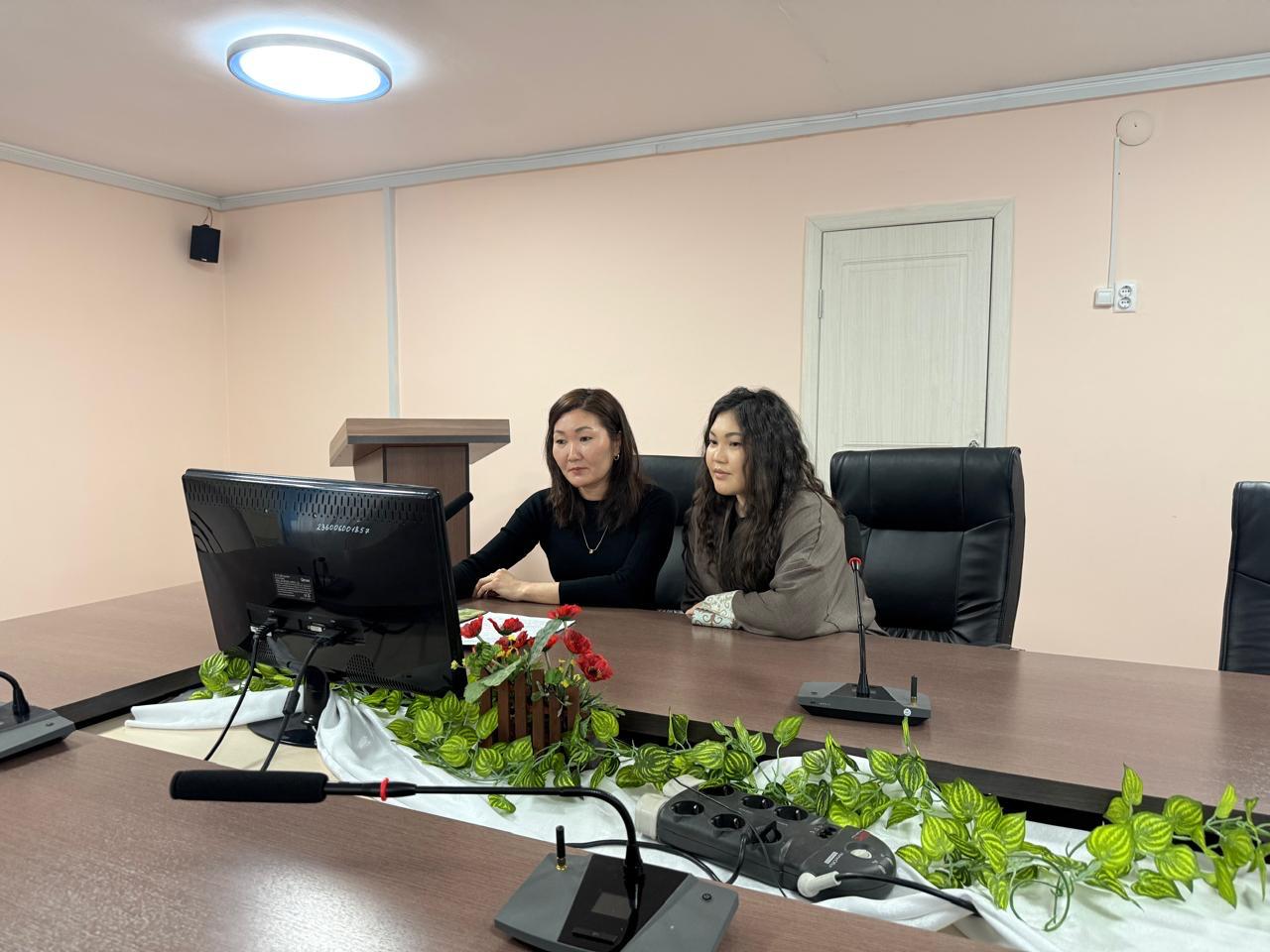Кеңседегі үстел басында отырған екі әйел. Үстелде монитор, микрофондар мен гүлдер бар.