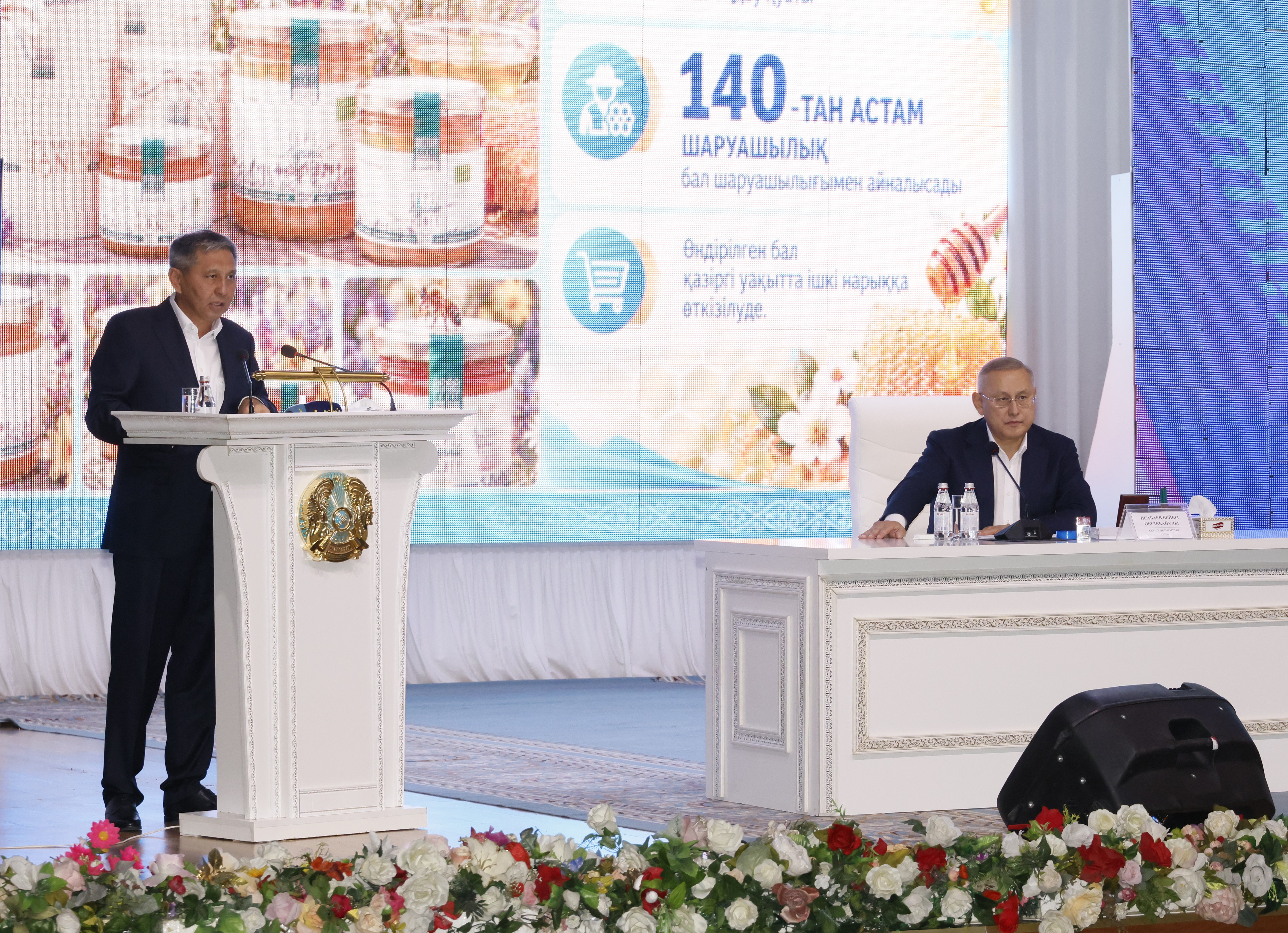 Сөйлейтін трибунадағы спикер және үстелде отырған адам.